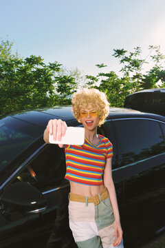 Woman with curly blond hair wearing a striped crop top and denim skirt, posing by a sleek black car, taking a selfie under bright daylight, retro film color mood.
