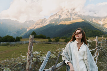 Fototapeta premium Cheerful woman outdoors wearing cozy cardigan near a rustic fence, mountain backdrop, soft sunlight, relaxed lifestyle moment, natural tones, lush landscape