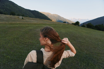 Fototapeta premium Young woman with a backpack stands in a expansive green field, pulling her long hair back as distant mountains glow in the late light, conveying freedom