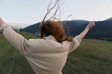 Fototapeta premium Woman in beige jacket raises arms in a windy countryside, embracing nature and freedom, a wide meadow and distant mountains under a bright sky, a pure joyful outdoor moment