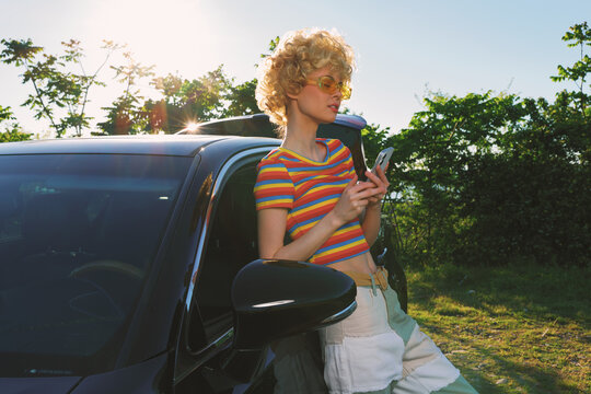 A woman leans against a dark car in a sunny outdoor setting, wearing a striped tee and sunglasses. A retro color palette enhances a casual lifestyle moment.