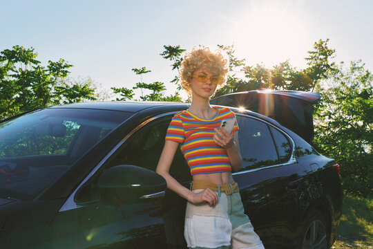Smiling woman beside a car wearing a striped crop top in bright sunlight, outdoor scene conveying a casual lifestyle, summer vibe, and vintage film warmth, under soft natural lighting.