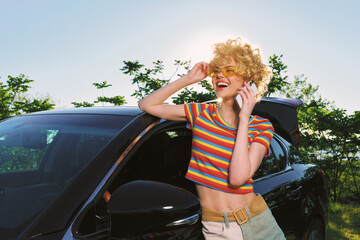 Young woman with curly blonde hair in a striped crop top talks on a mobile phone beside a dark car...