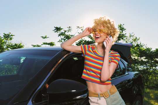 Young woman with curly blonde hair in a striped crop top talks on a mobile phone beside a dark car in a sunny outdoor setting, exhibiting a cheerful mood and casual vibe