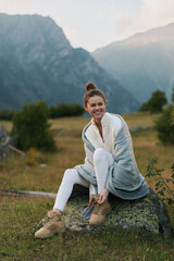 Fototapeta premium Young woman sits on a mossy rock in a mountain meadow, adjusting her hiking boot and smiling softly as warm light highlights her relaxed, hopeful mood.