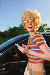 A cheerful woman with retro blond curls and yellow sunglasses stands beside a sleek car in bright...