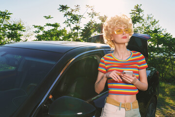 Fototapeta premium Woman outdoors, retro fashion, striped crop top, standing beside a car in bright sunlight, casual lifestyle, vintage colors, lush green background, carefree moment