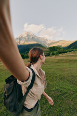 Fototapeta premium A young woman with a backpack walks across a green meadow toward distant mountains, pausing to admire rugged peaks and breathe in the fresh air.