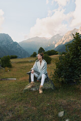 Fototapeta premium A woman sits on a rock in a mountain meadow, wrapped in a soft poncho, enjoying a quiet moment in nature. A serene lifestyle scene of relaxation and exploration.