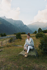 Fototapeta premium A woman sits on a rock in a scenic meadow with distant mountains, conveying relaxed outdoors lifestyle and peaceful solitude in nature with light breeze, and open sky.