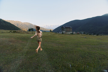 Fototapeta premium Young woman runs barefoot across a green valley with distant mountains and a rustic house, capturing a sense of freedom and carefree outdoor lifestyle in warm afternoon light.