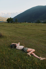 Fototapeta premium Young woman lying in a grassy field near mountains, enjoying outdoor relaxation, casual clothing, peaceful mood, nature escape, sunlit landscape under a clear blue sky
