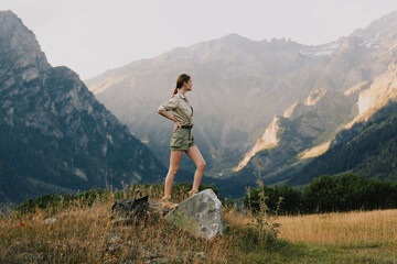 Fototapeta premium Young woman in hiking shorts and beige top stands on a rock amid a rugged mountain valley, enjoying a moment of exploration and freedom with a sense of adventure and calm as the wind sweeps the grass.