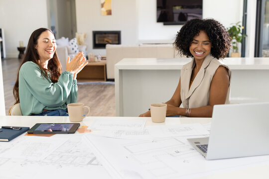 Diverse female colleagues reviewing design blueprints on kitchen island, using laptop, tablet