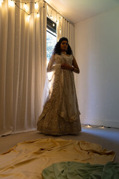 Indian woman wearing ornate beaded gown and sheer dupatta, standing by window with fairy lights