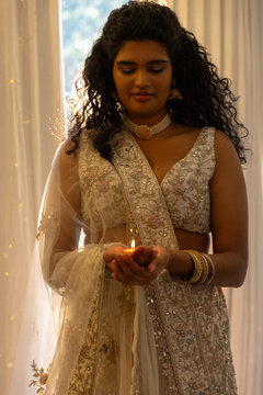 Indian woman standing centered in softly lit room holding lit diya wearing embroidered lehenga