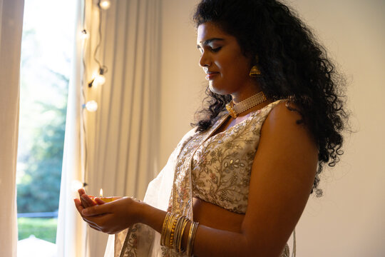 Adult Indian woman holding lit diya, gazing by window at home with fairy lights, wearing jhumka