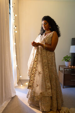 Indian woman standing near tall window with sheer curtains and fairy lights, wearing ornate lehenga
