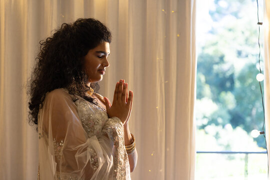 Indian woman pressing palms, standing by window, wearing sheer dupatta and gold bangles, copy space