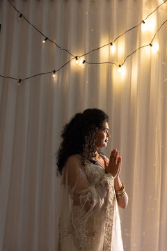 Indian woman standing in profile pressing palms at chest in warm room with dupatta, bangles, lights