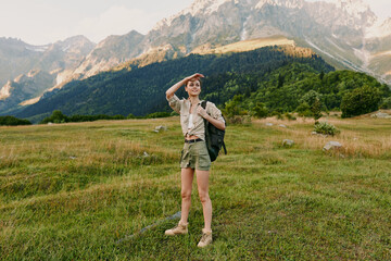 Fototapeta premium Woman with backpack standing in a grassy meadow near mountains, ready for adventure. Outdoor lifestyle scene with natural light and expansive landscape.