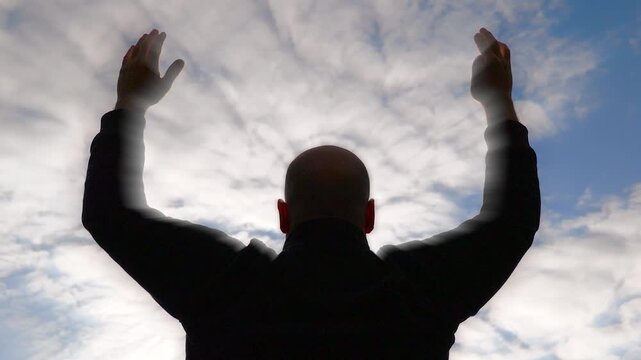 Man with both arms and hands up, slowly lowering head   and bowing at the sky. Strong light and sky full of clouds in the background.