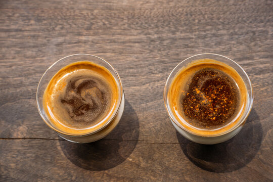 Two cups of coffee with latte art on a wooden table. The left cup features a heart-shaped design, while the right cup displays a circular pattern. The focus is on the intricate details of the latte