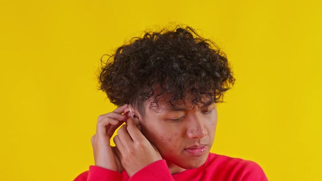 Young Asian Man Putting On Hearing Aid Against Bright Yellow Background