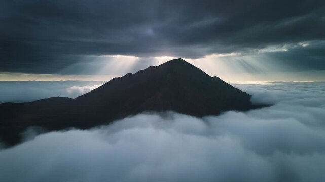 Volcanic mountain peak rising above a dense sea of white clouds under dramatic storm clouds with sun rays.