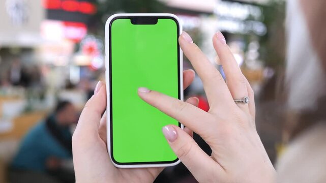 Hand holds phone with green screen. Background blurred indoor public area. Fingers interact with device. Person wears light-colored clothing. Scene shows casual usage
