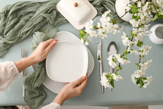 Woman setting table with beautiful blossoming branches, plates, glass and fabric in dining room