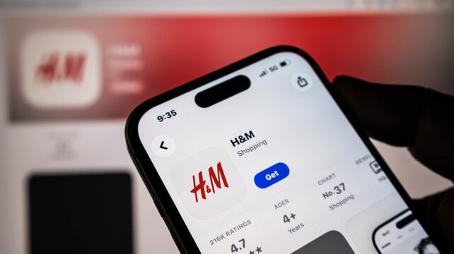 Closeup of a smartphone screen displaying the hm shopping app on the apple app store, held in a hand with a blurred background