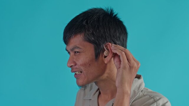 Side Profile Of Smiling Asian Man Adjusting Behind-The-Ear Hearing Aid On Blue Background