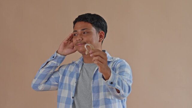Young Asian Man Holding Hearing Aid And Cupping Ear To Listen Attentively On Beige Studio Background