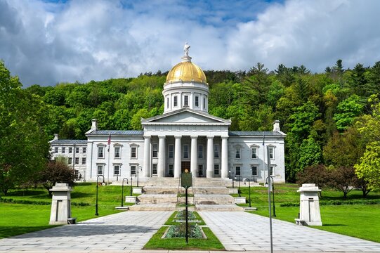Front view of the Vermont State House in Montpelier, featuring a gold dome and neoclassical architecture, surrounded by lush green trees on a bright spring day