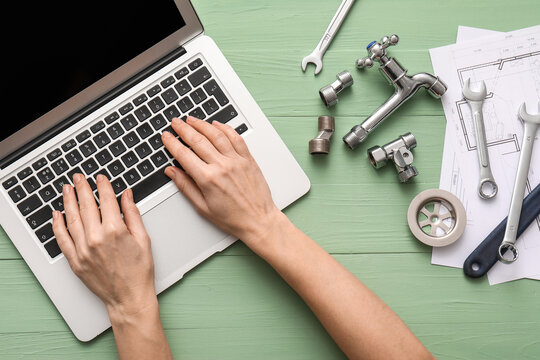 Female hands with laptop, set of plumber's items and house plans on green wooden background