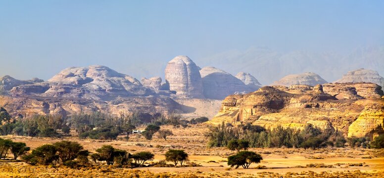 Scenic desert landscape of Jabal Ithlib and the ancient rock formations of Hegra, Al Ula, Saudi Arabia, featuring sandstone mountains and scattered greenery under a clear sky.