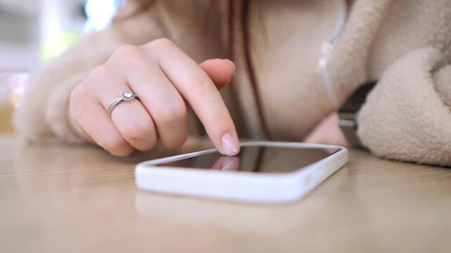 Close-up of a woman's hand tapping a smartphone. Fingers press screen with ring visible. Hand rests on wooden table surface. Soft lighting highlights casual indoor setting