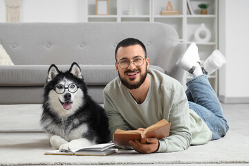 Handsome young man lying on floor and reading book with funny husky dog in eyeglasses at home © Pixel-Shot