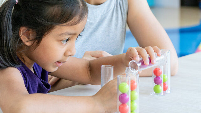 Happy Asian mother watching her daughter playing educational sorting game with test tubes, spending quality family time together in living room.
