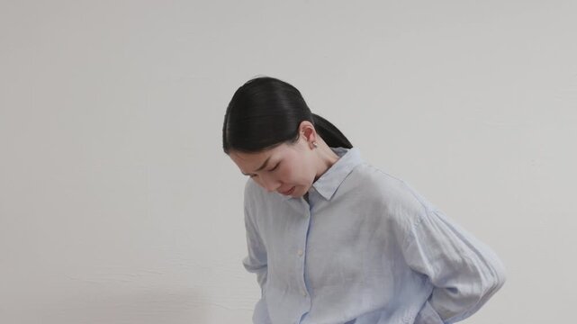 Asian Woman Touching Lower Back with Painful Expression in Front of White Wall