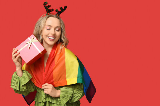 Beautiful young woman in reindeer horns with LGBT flag and gift box on red background. Christmas celebration