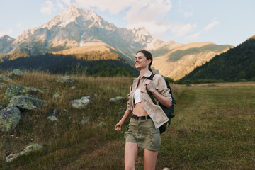 Fototapeta premium A woman hiker walks through a bright meadow toward distant mountains, wearing shorts, a light shirt, and a backpack. Concept: outdoor exploration and active lifestyle.