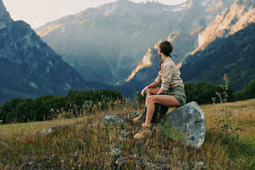 Fototapeta premium Bright outdoor lifestyle scene with a woman sitting on a rock in a meadow, mountains in the background, relaxed pose, natural light, gentle breeze, warm tones