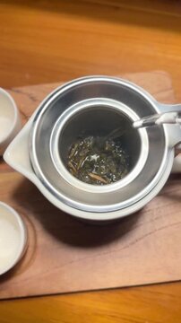 Water pours over dark tea leaves in a ceramic strainer. Hand gently presses the strainer to release tea. Two small bowls sit nearby on a wooden board. Warm lighting highlights the tea-making process