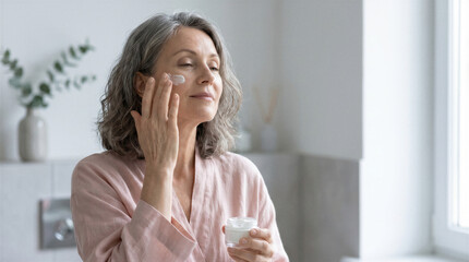 Fototapeta premium Middleaged woman with gray hair applying moisturizing cream on her face in the bathroom, skincare routine and selfcare concept