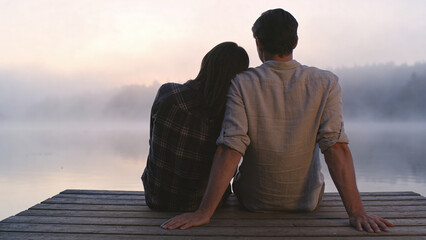 Obraz premium Couple sitting on a wooden dock overlooking a misty lake at sunrise