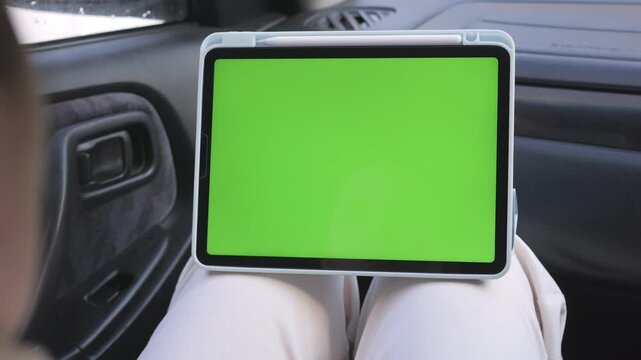Person holds tablet with green screen on lap inside car. Car interior visible. No animals or plants shown