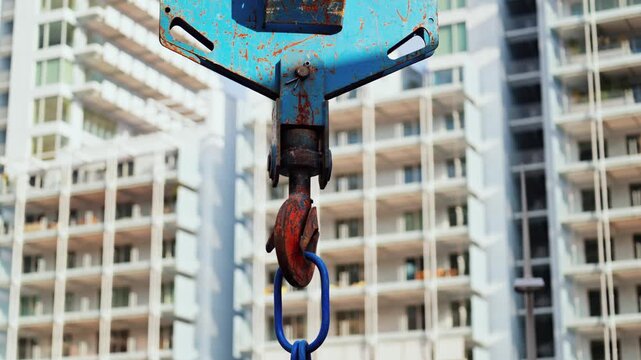 Industrial crane hook hanging with steel chain against modern residential apartment building construction background.