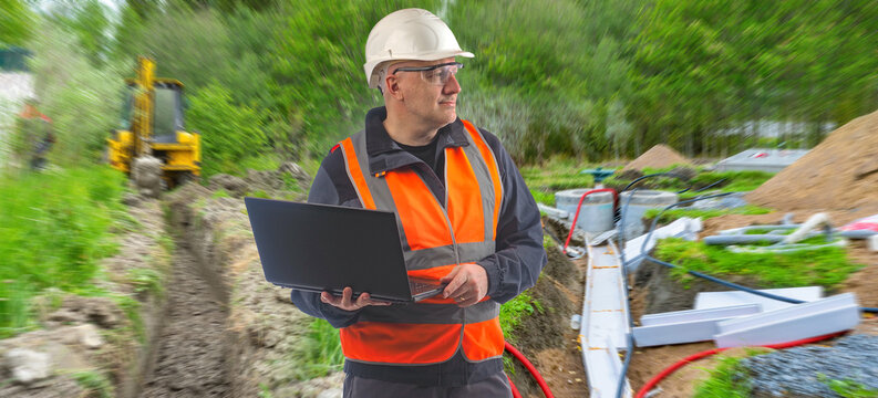 Utility construction, Infrastructure monitoring. Engineer checks trench works and underground cable installation with laptop at outdoor construction site.